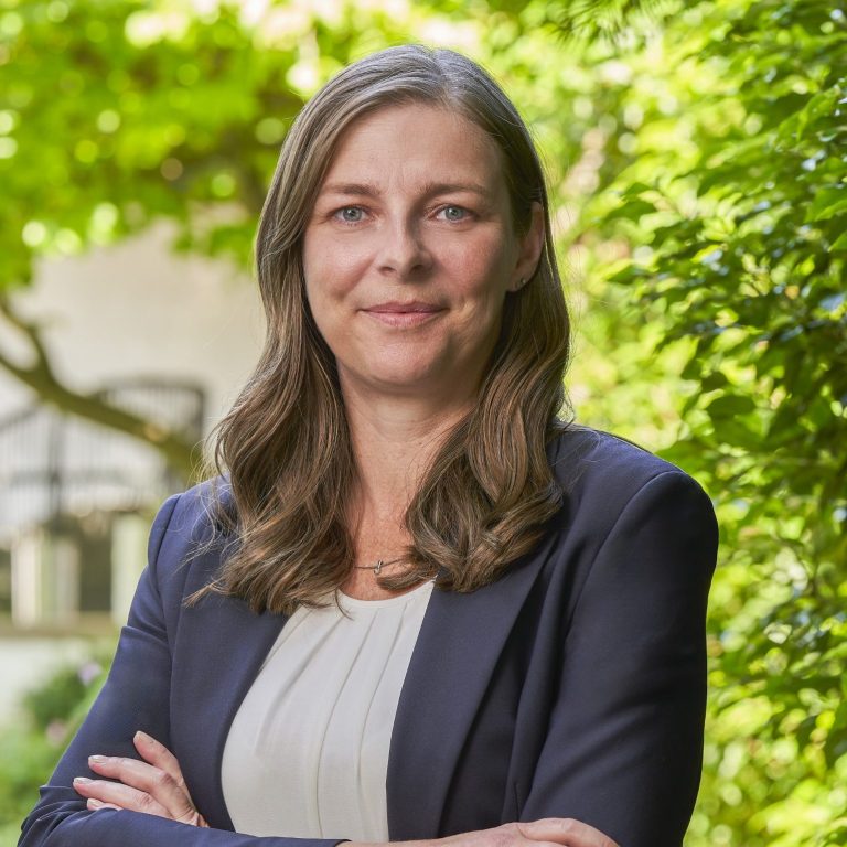 Dipl.-Kffr. Maike Thielmann Frau mit langen Haaren, in formeller Kleidung, steht vor grüner Pflanze und lächelt.