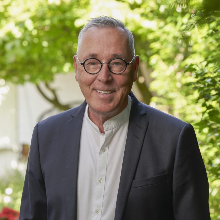 Prof. Dr. Manfred Hunkemöller Mann mit Brille und Anzug, steht lächelnd vor grüner Gartenlandschaft.